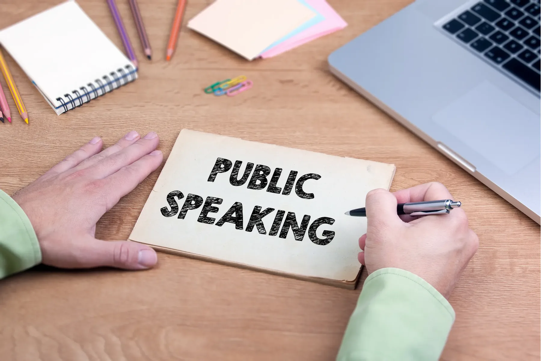 Hands writing the words public speaking on a card at a wooden desk, with a notebook, coloured pencils and a laptop.