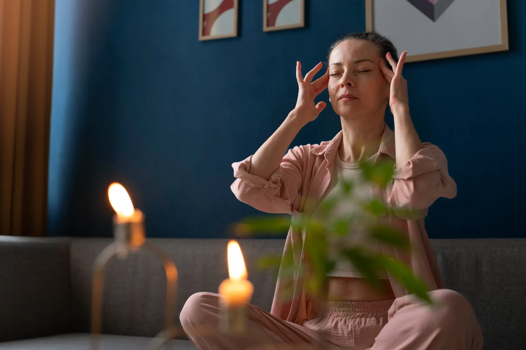 A woman sitting cross-legged on a sofa with her eyes closed and fingertips resting at her temples, in a calm candle-lit room.