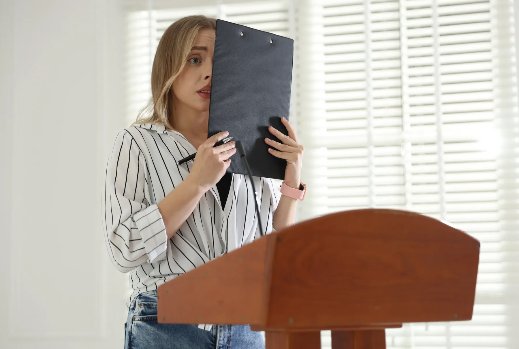 A speaker at a wooden lectern holding a clipboard in front of her, looking anxious before beginning to speak.