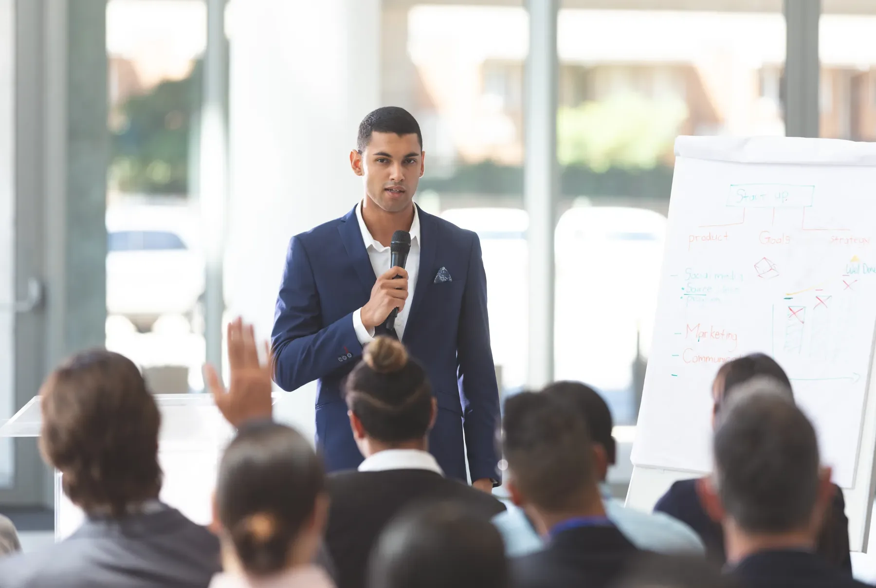 A man in a suit presenting to a seated audience with a flip chart of business notes beside him.
